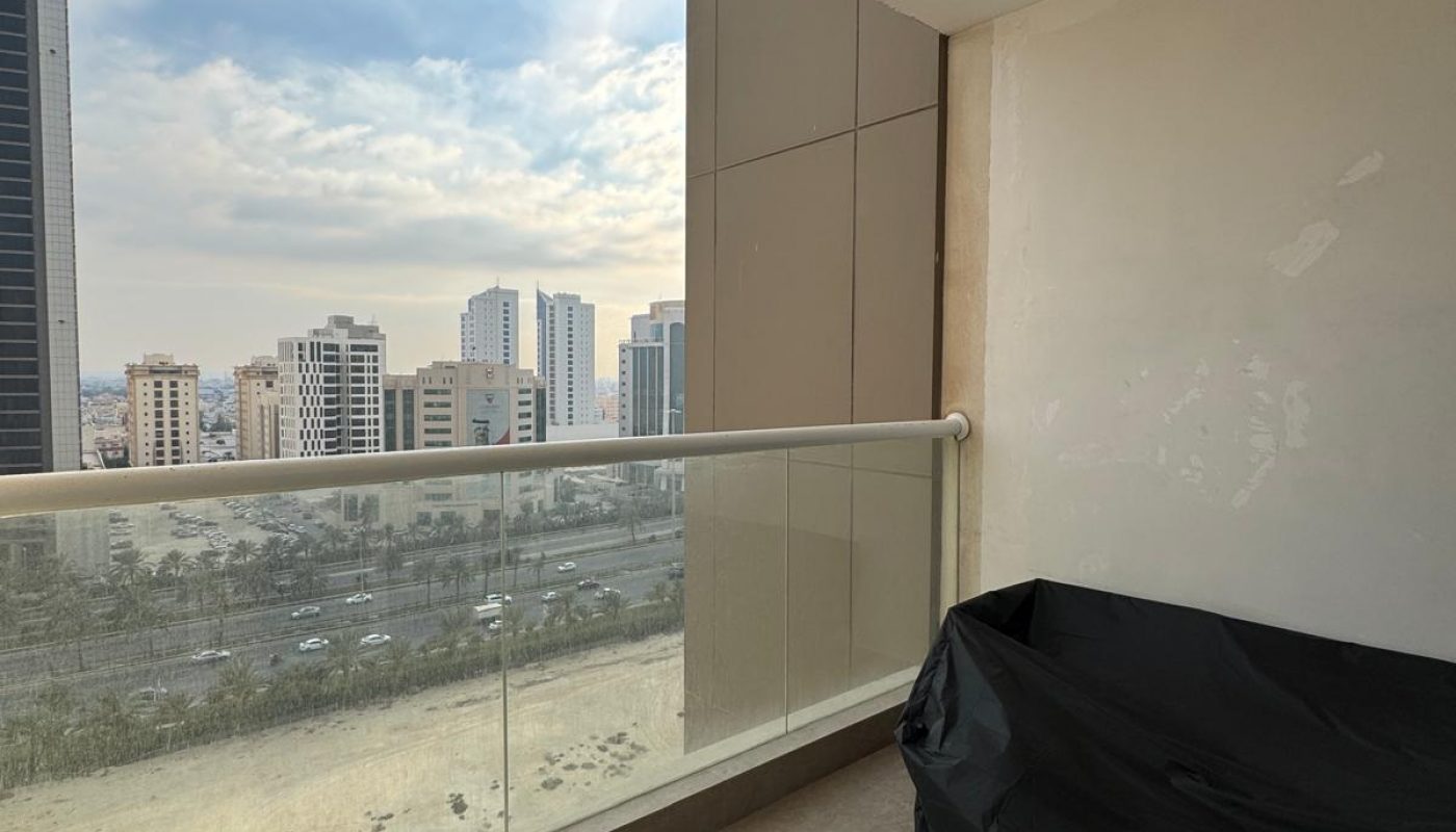 A high-rise balcony with a glass railing, tiled floor, covered item on the right, and a cityscape view of buildings under a partly cloudy sky.