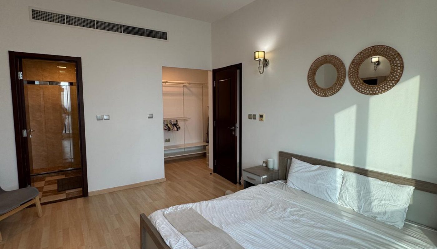 A modern bedroom with a neatly made double bed, wooden flooring, wall-mounted lamps, two round decorative mirrors, and an open closet visible through a doorway.