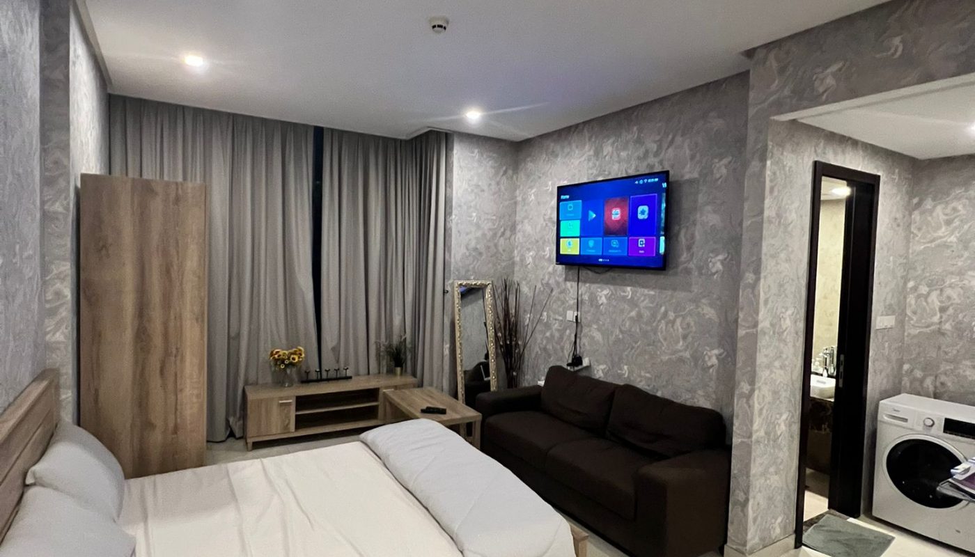 Modern bedroom with a double bed, brown sofa, wall-mounted TV, wardrobe, and a visible washing machine in the adjacent bathroom area. Neutral colors and patterned wallpaper.