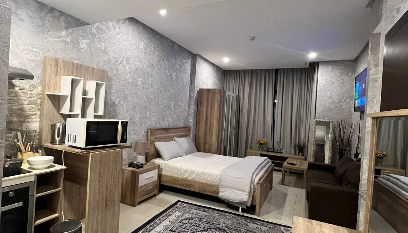 A modern studio apartment with a bed, kitchenette, microwave, TV, brown sofa, and a large patterned rug, featuring neutral tones and floor-to-ceiling curtains.