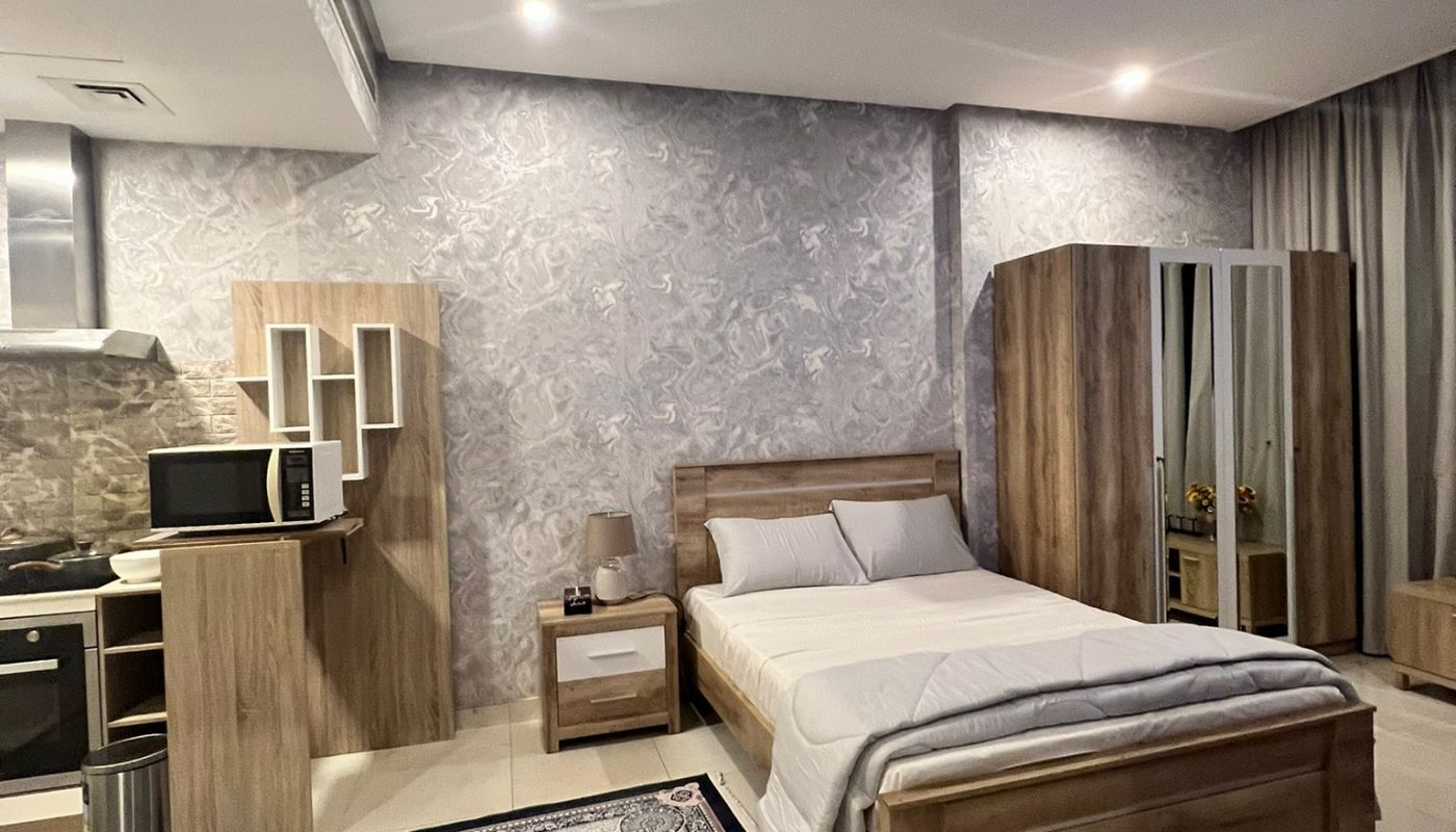 A modern bedroom features a double bed, bedside table, wardrobe, kitchen area with appliances, and a patterned rug on a tiled floor.
