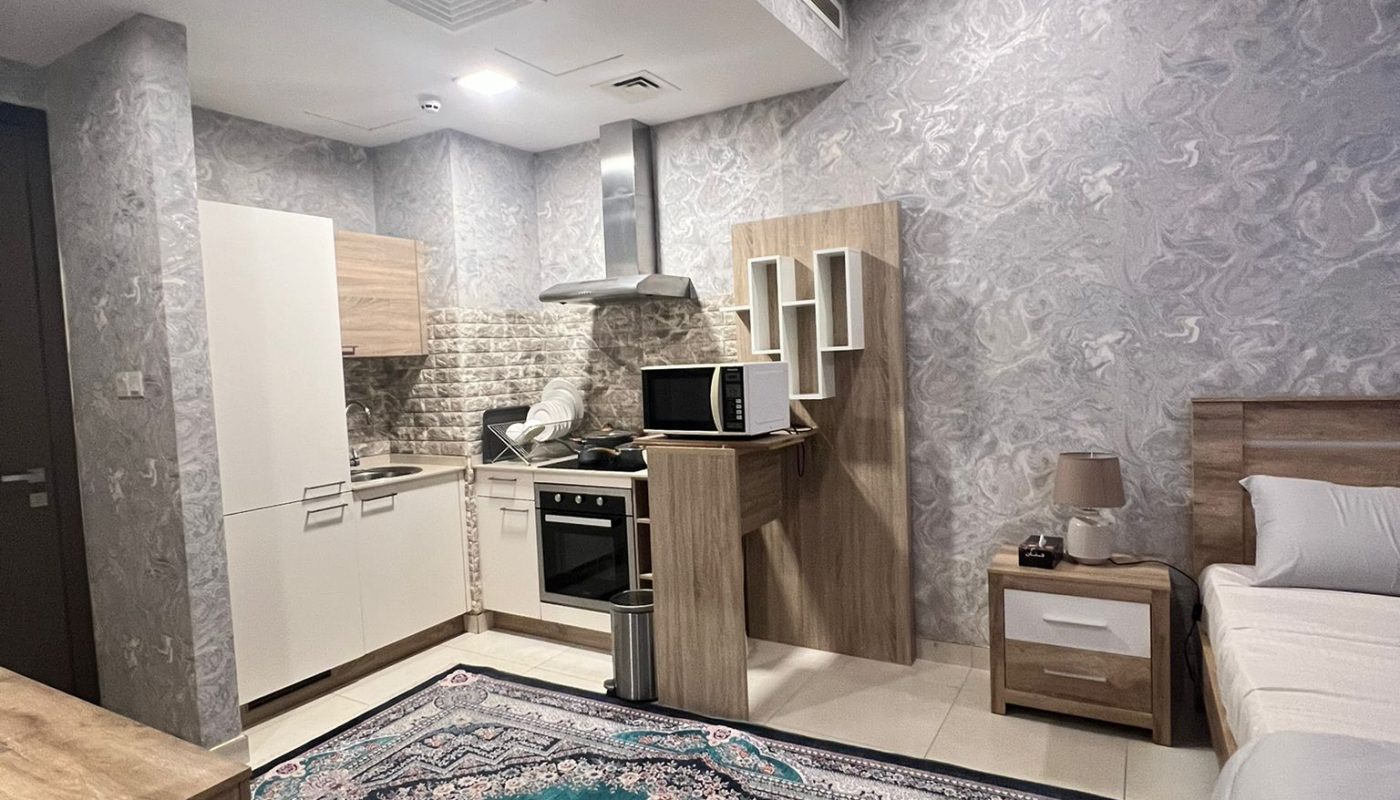 A modern studio apartment with a kitchen, a single bed, a nightstand with a lamp, and a large patterned rug on the floor.