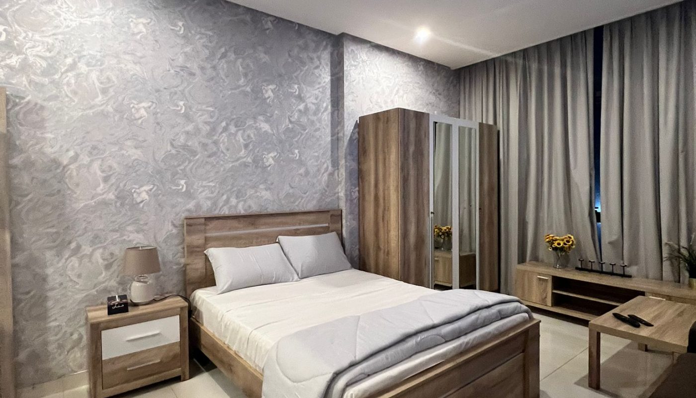 A modern bedroom with a double bed, wooden furniture, patterned rug, bedside table with lamp, wardrobe, and floor-to-ceiling curtains.