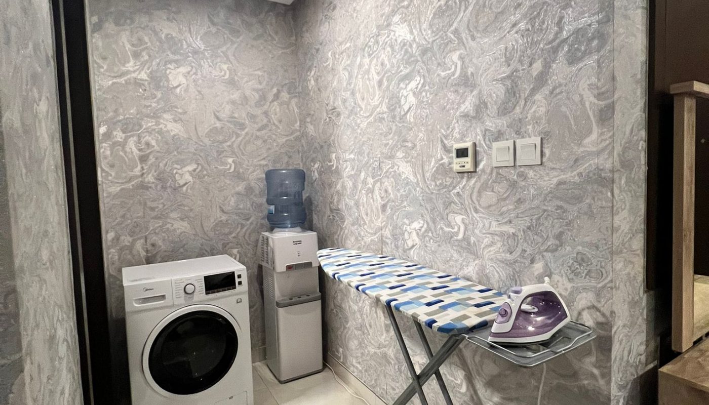 A laundry area with a front-load washing machine, a water dispenser, an ironing board with an iron, and a gray floor mat on tiled flooring.