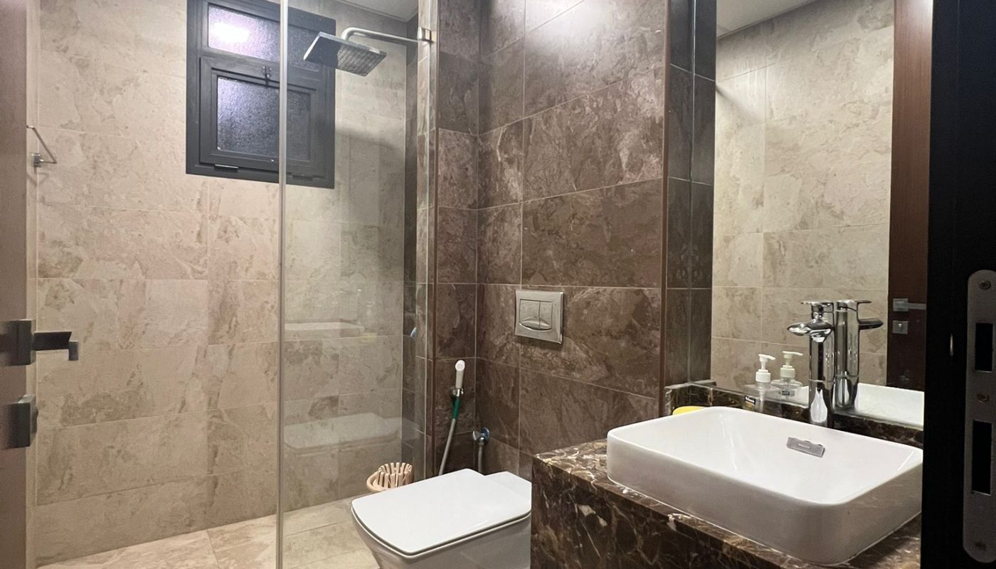 Modern bathroom with marble walls and floor, a walk-in shower, wall-mounted toilet, large sink with countertop, mirror, and various toiletries on the counter.