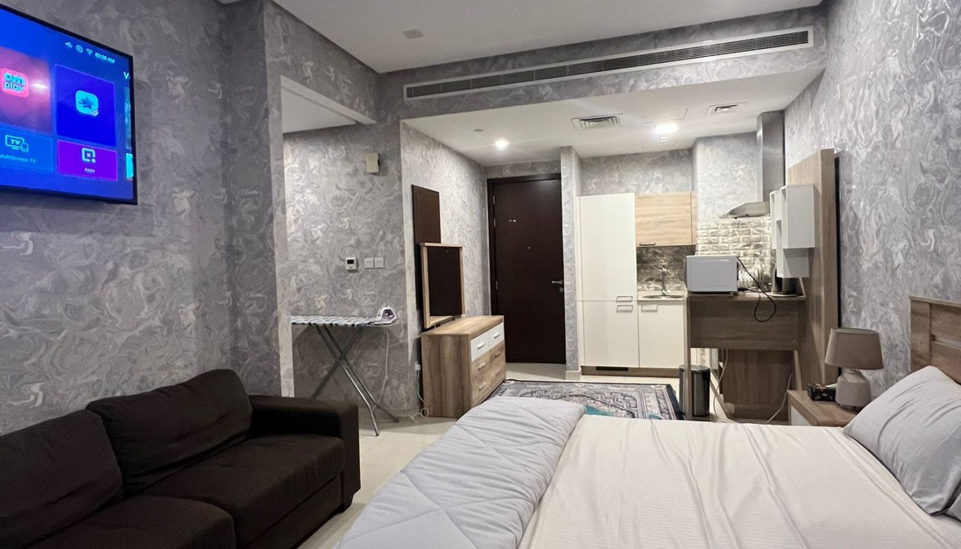 A modern studio apartment with a double bed, brown sofa, wall-mounted TV, kitchenette, and built-in wardrobe, decorated in neutral tones with tiled flooring.