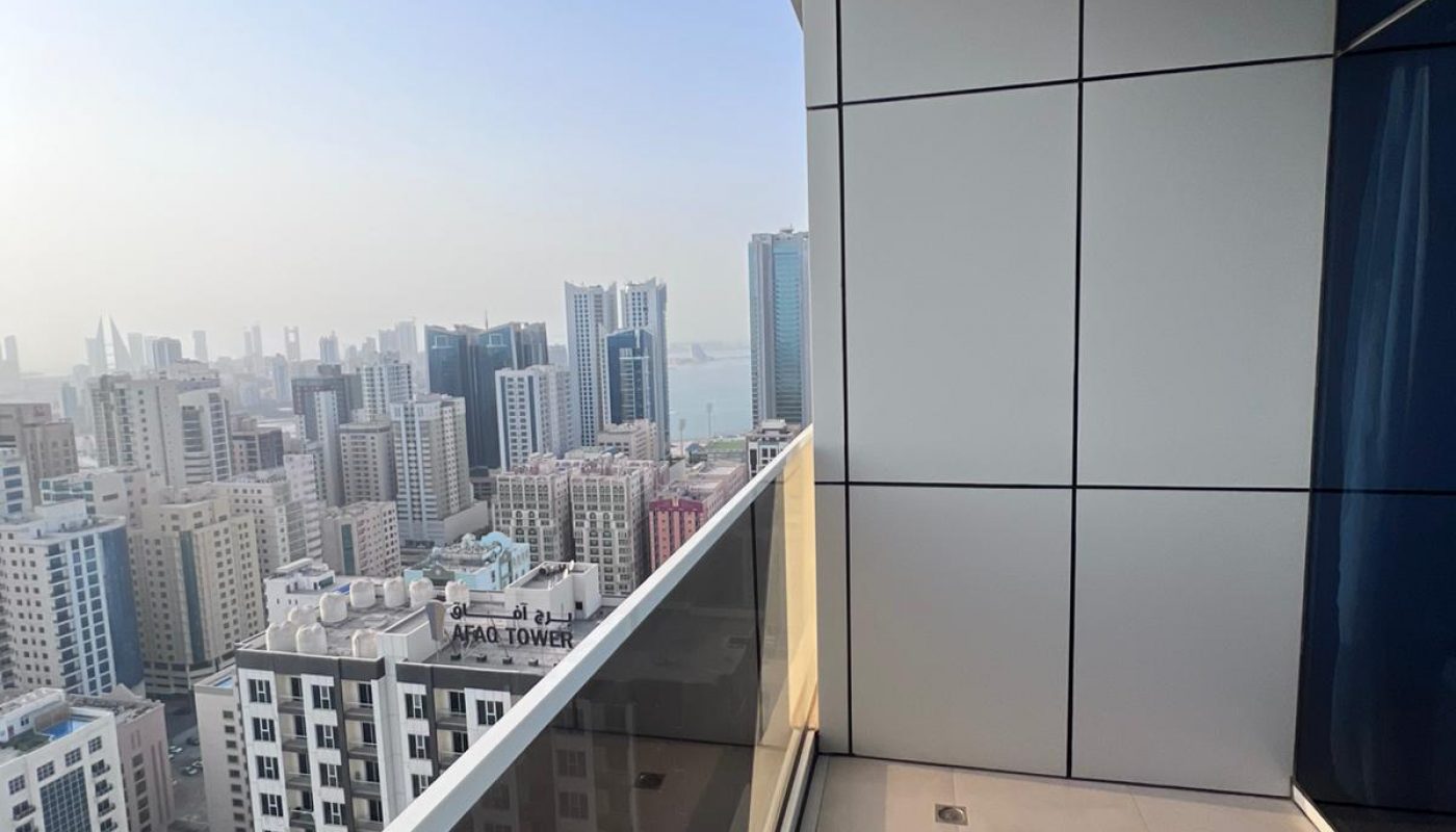 View from a high-rise balcony overlooking a cityscape with tall buildings, modern architecture, and a distant body of water under a clear sky.