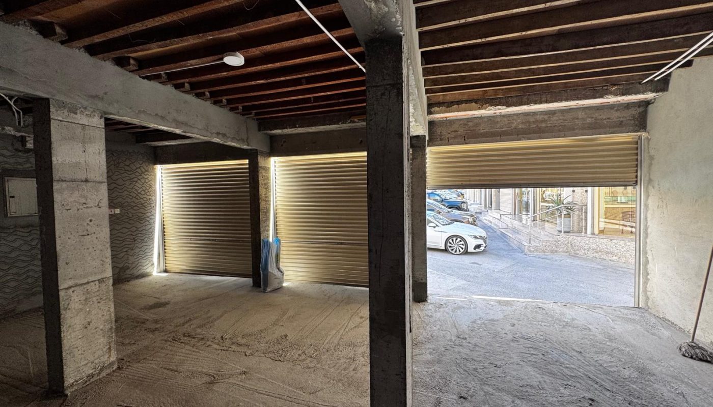 Interior view of an unfinished garage with open and partially closed metal roll-up doors, exposed ceiling beams, and a parked car visible outside.