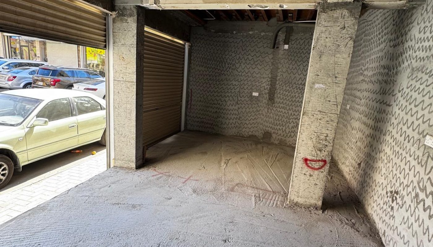 A small, empty garage with unfinished concrete floor and exposed ceiling beams; garage door is open, and two parked cars are visible outside.