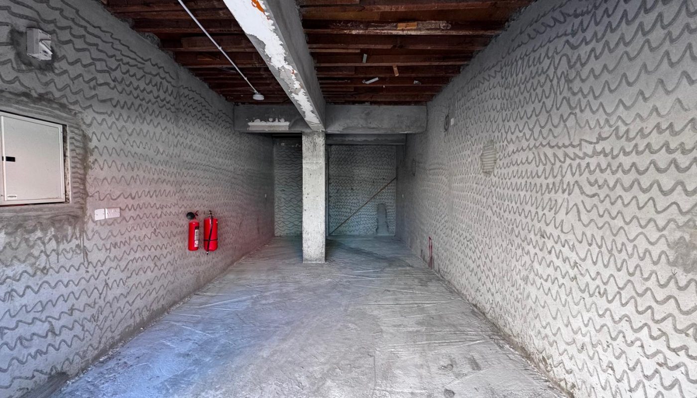 Unfinished empty room with concrete floor and walls, exposed ceiling beams, a central support column, and two red fire extinguishers mounted on the left wall.