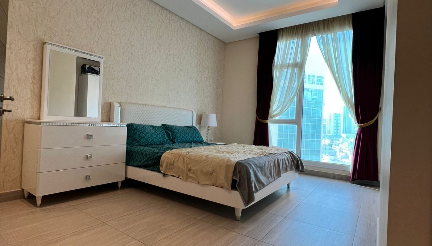 Modern bedroom with a double bed, two nightstands, a dresser with a mirror, large window with curtains, and cityscape view. The room has beige tiled flooring and recessed ceiling lighting.