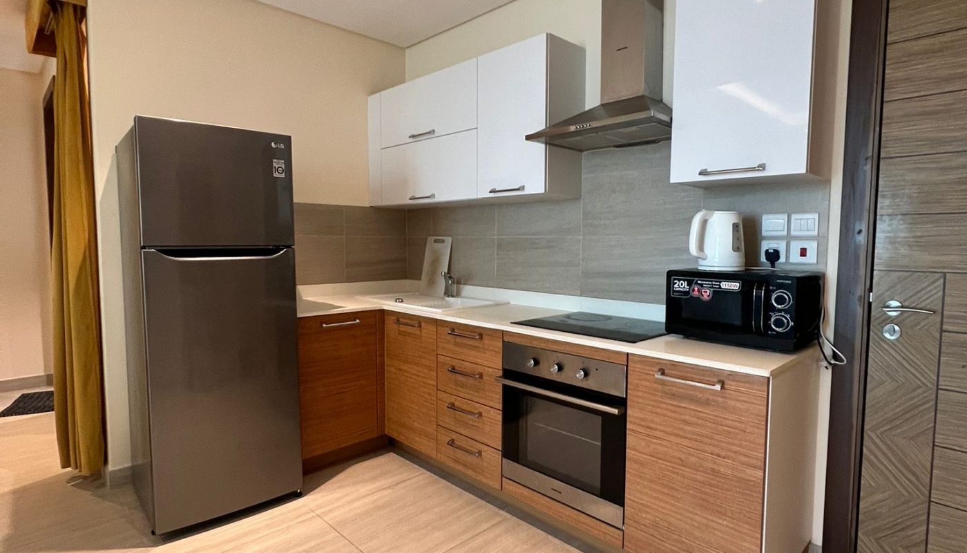 Modern kitchen with wooden cabinets, stainless steel appliances including a fridge, oven, microwave, and kettle, beige tiled floor, and white upper cabinets.
