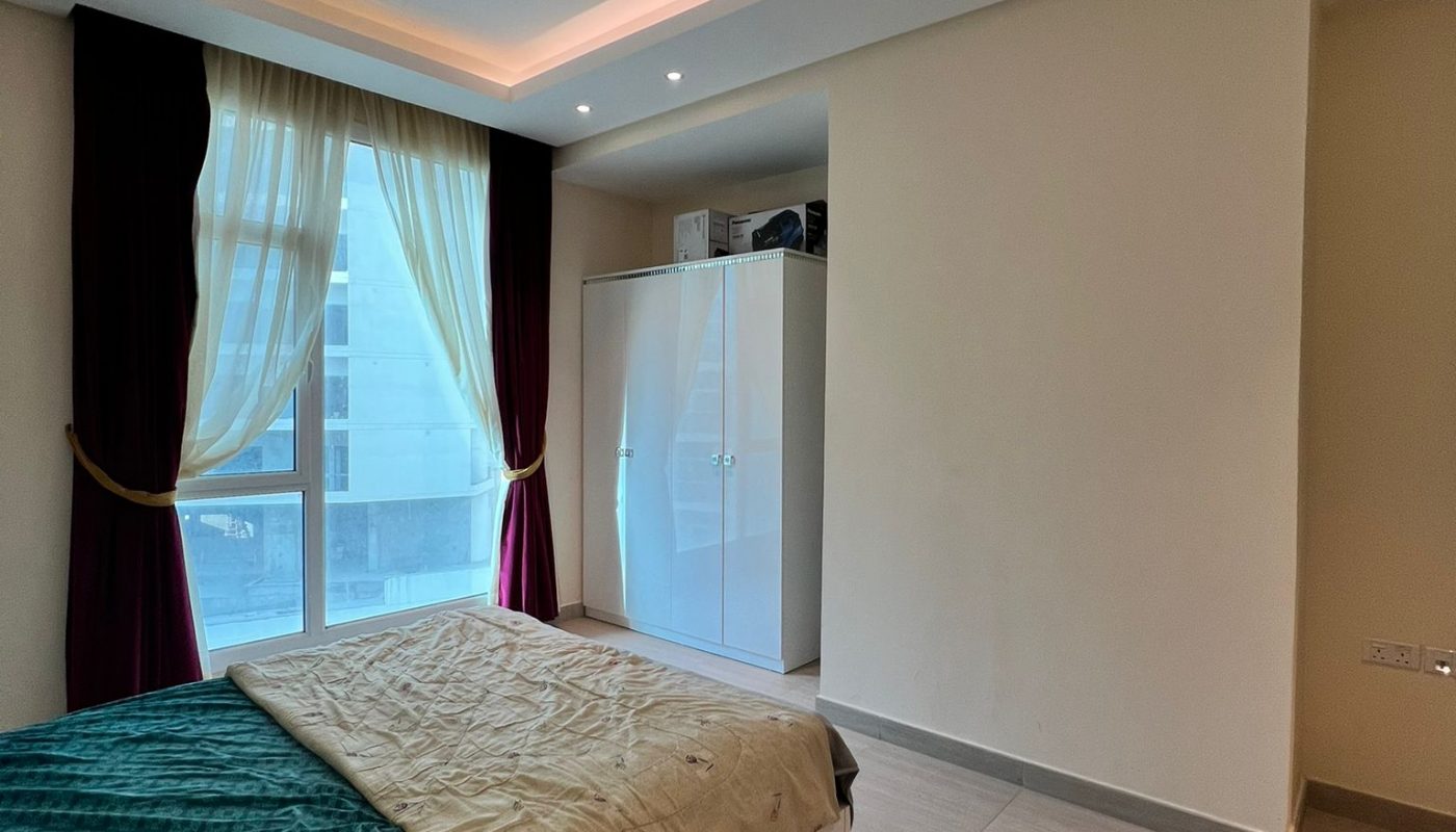 A modern bedroom with a low bed, teal and beige bedding, a large window with curtains, a white wardrobe, and neutral tile flooring.
