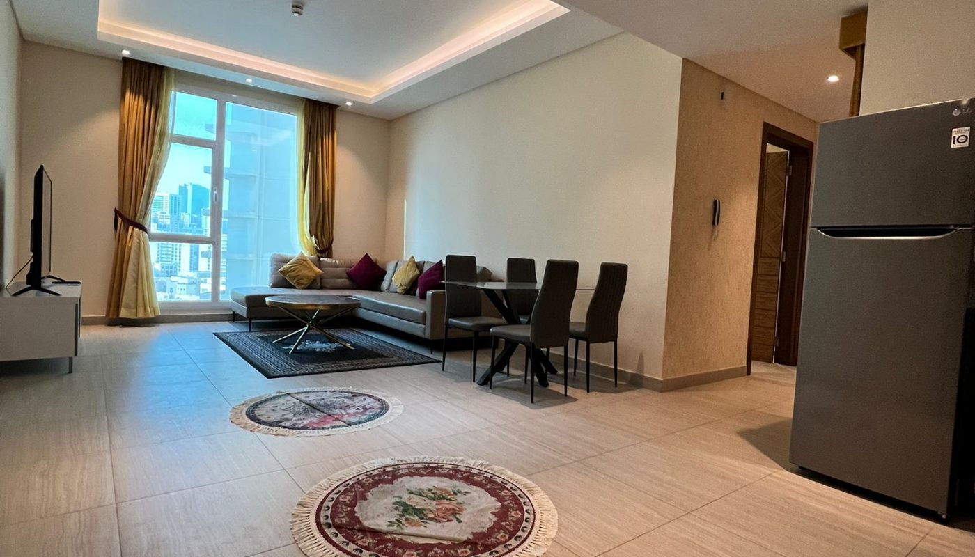 A modern living and dining area with tiled floors, a gray sofa, a dining table with four chairs, two round rugs, a TV, and a refrigerator, with natural light from a balcony window.