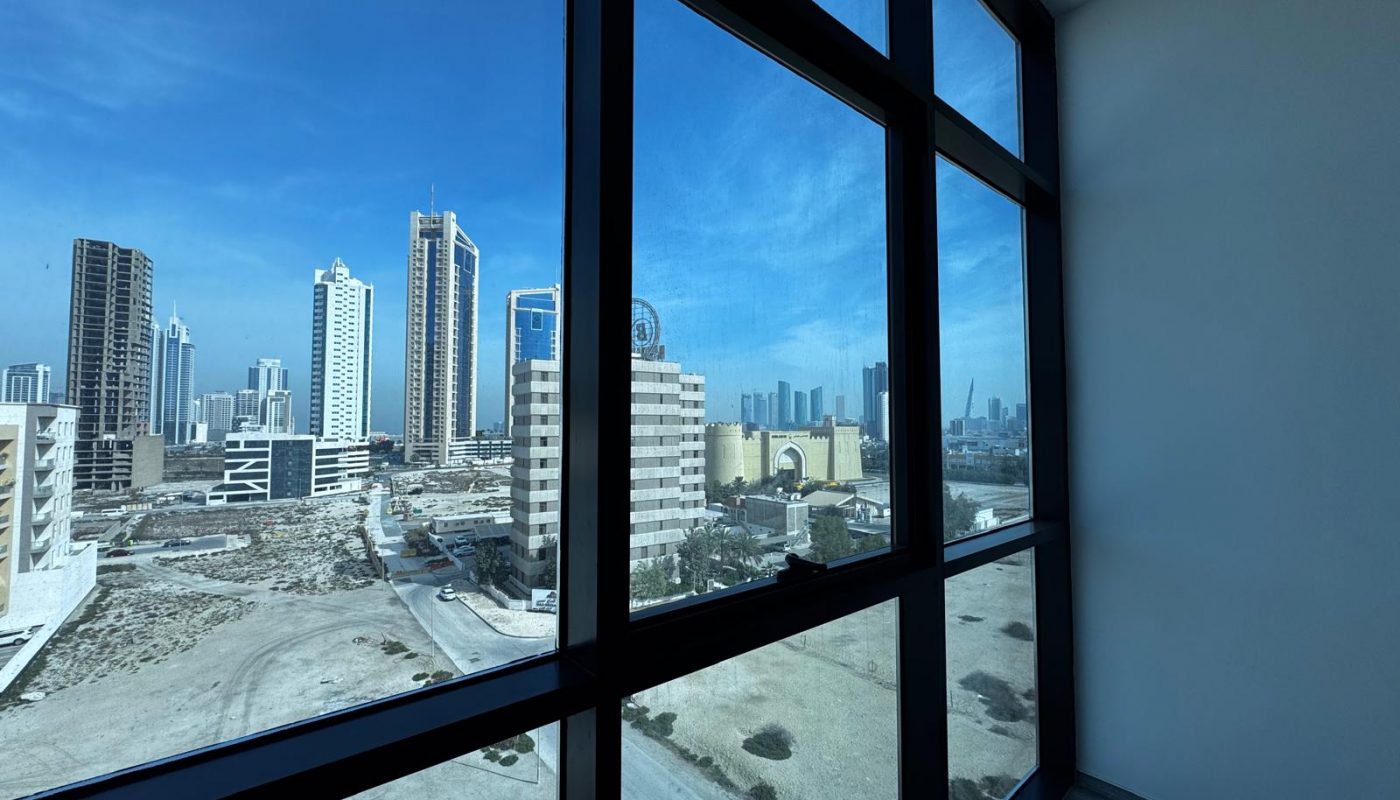 Cityscape view through large glass windows from a high-rise building, showing modern skyscrapers and empty lots under a clear blue sky.