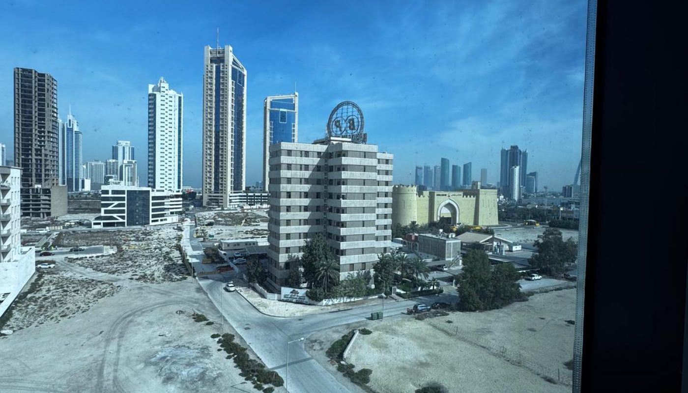 View from a window overlooking a cityscape with tall buildings, a few parked cars, sandy lots, and a partly cloudy blue sky.