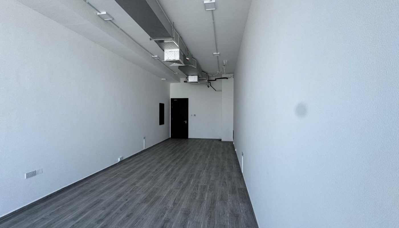Empty rectangular room with white walls, gray wood flooring, exposed ceiling ducts, and a black door at the far end.