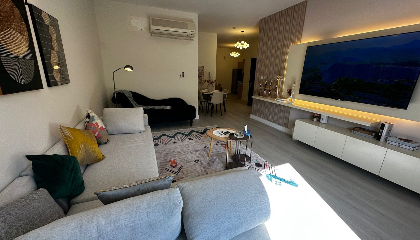 Modern living room with light gray sectional sofa, wall-mounted TV, contemporary lighting, and dining area in the background. Sunlight streams through large windows onto the floor.