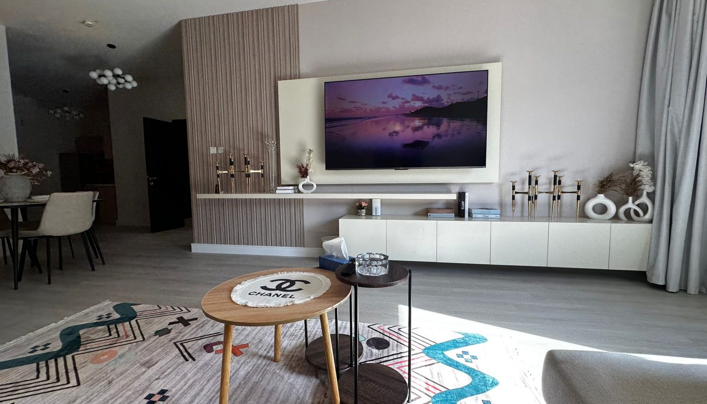 Modern living room with a wall-mounted TV displaying a landscape, light wood furniture, decorative items, and a patterned rug; sunlight streams through large windows.