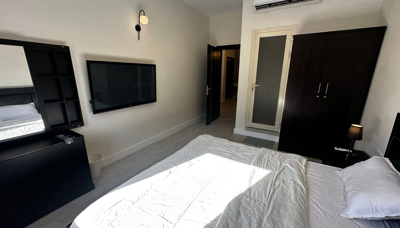 A modern bedroom with a made bed, wardrobe, wall-mounted TV, mirrors, air conditioner, and minimalistic lighting. Sunlight streams through a window onto the bed.