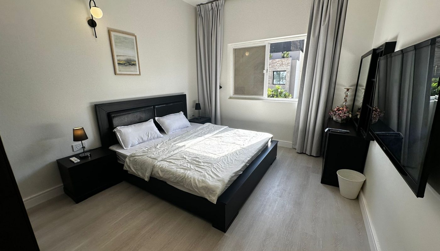 Modern bedroom with a black bed frame, white bedding, two bedside tables with lamps, wall art, large window with gray curtains, mounted TV, and light wood flooring.