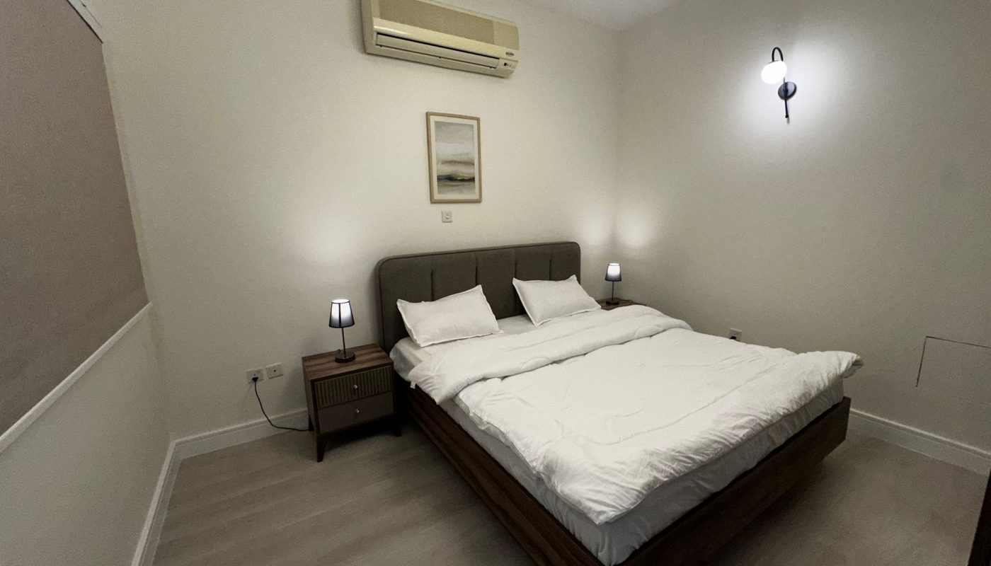 A minimalist bedroom with a double bed, two bedside tables with lamps, an air conditioner, a wall light, and a framed picture above the bed.