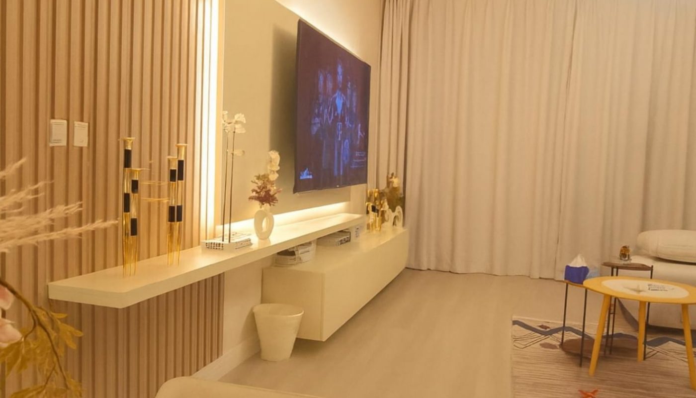 A modern living room with a wall-mounted TV, light wood paneling, white furniture, a patterned rug, and decorative items on shelves and table. The room is warmly lit with a ceiling light.