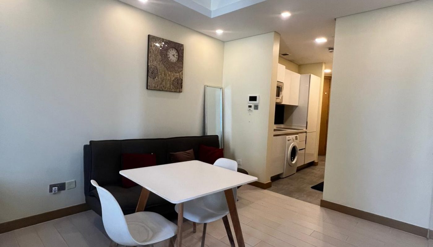 A small dining area with a white table and two chairs next to a black sofa, adjacent to an open kitchen with white cabinets and a washing machine.