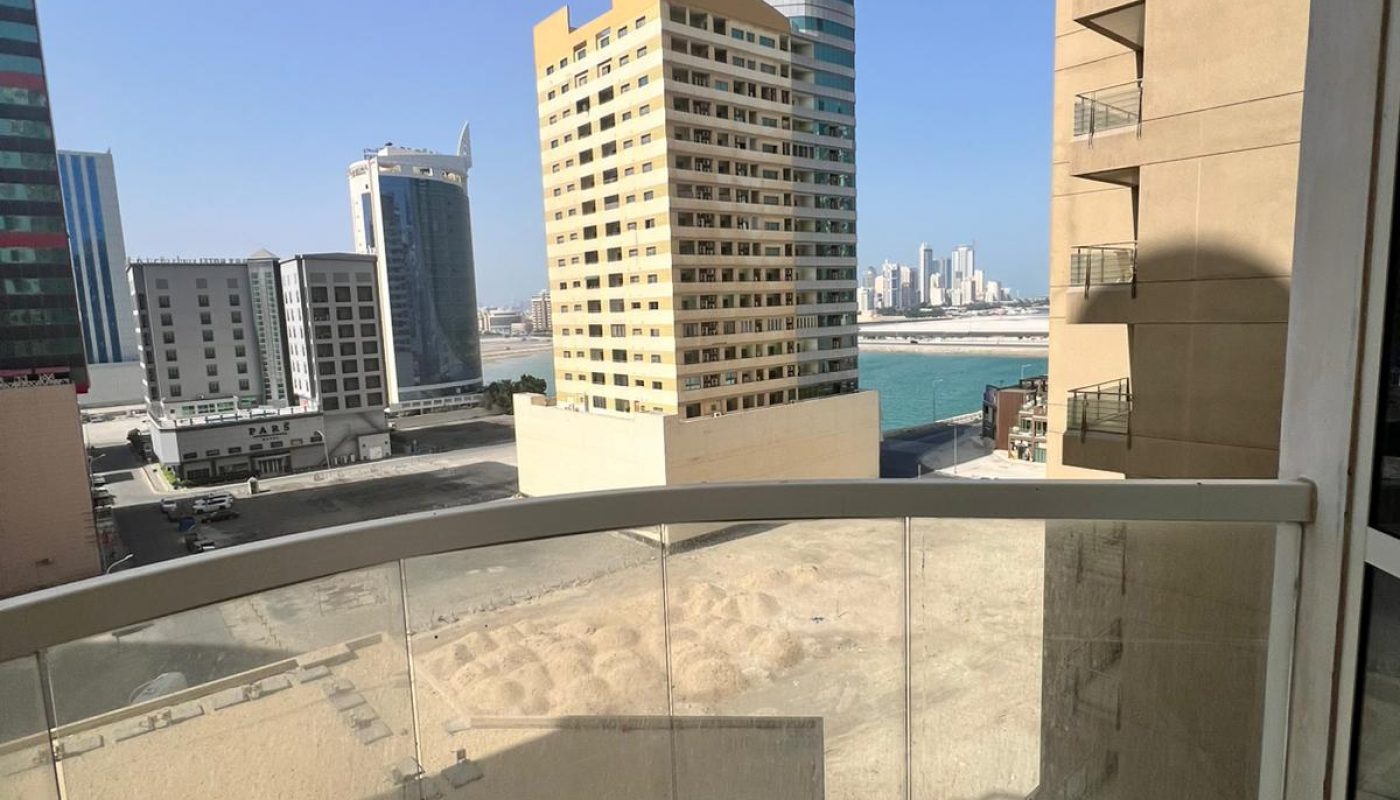 View from a balcony with glass railing overlooking a sandy lot and several modern high-rise buildings under a clear sky.