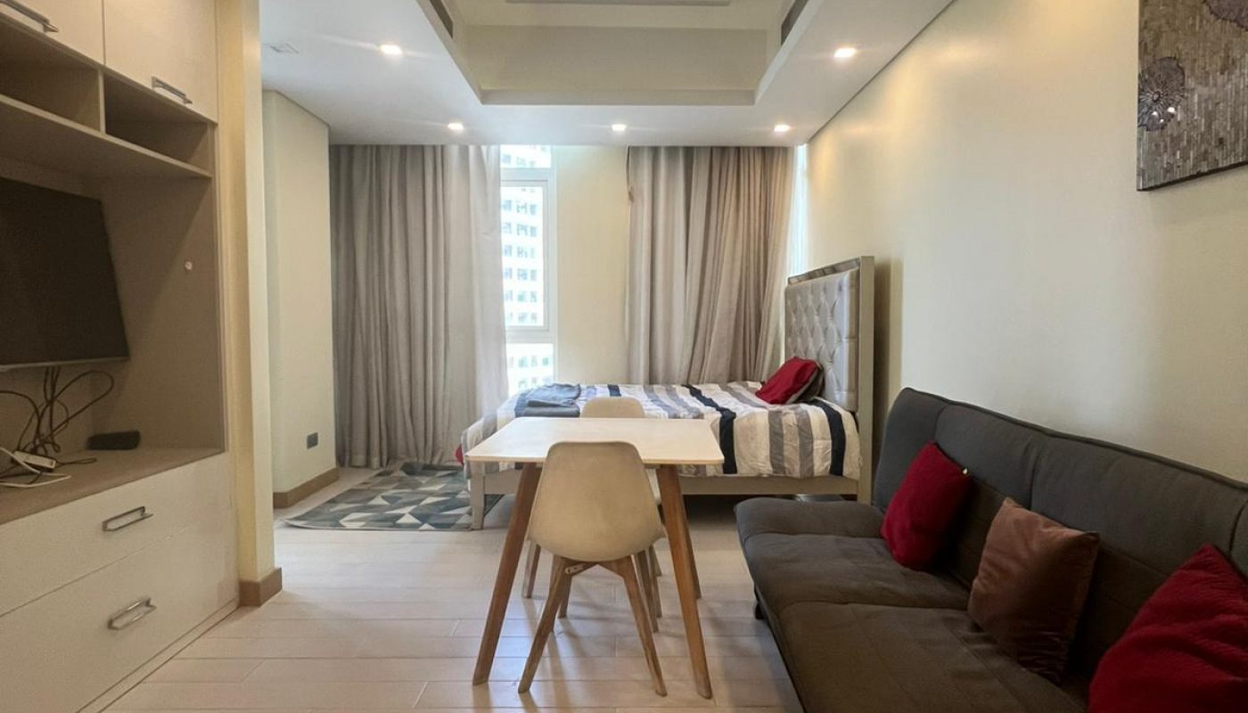 A modern studio apartment with a bed, sofa, small dining table, wall-mounted TV, shelves, and a window with curtains letting in natural light.