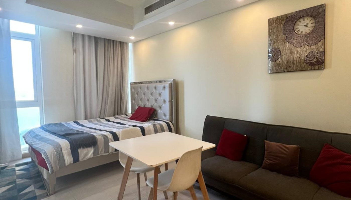 A modern studio apartment with a bed, small dining table with two chairs, dark sofa, and wall clock above, featuring light-colored flooring and large window with curtains.