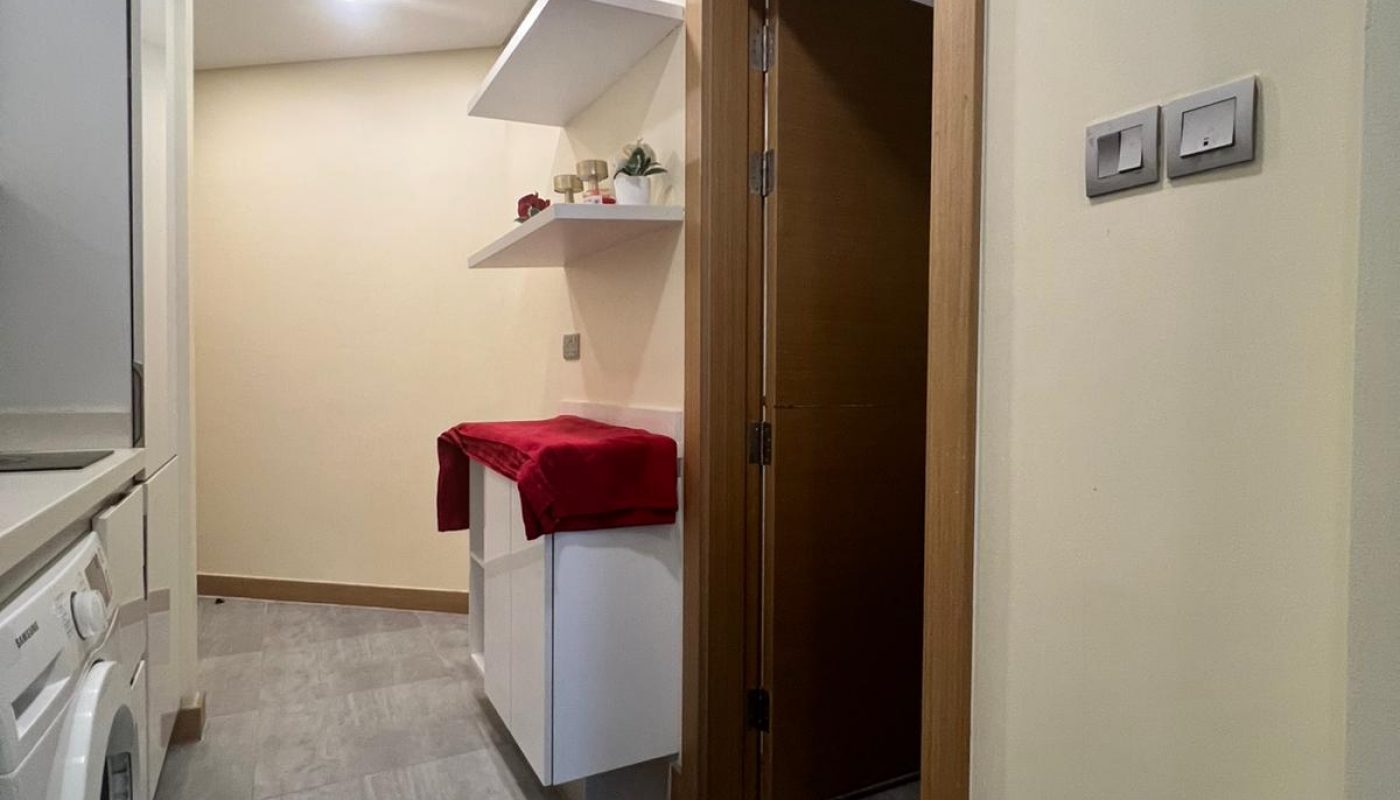 A laundry area with a washing machine, a cabinet covered with a red cloth, shelves with decor items, a closed wooden door, and a blue floor mat on gray tile flooring.