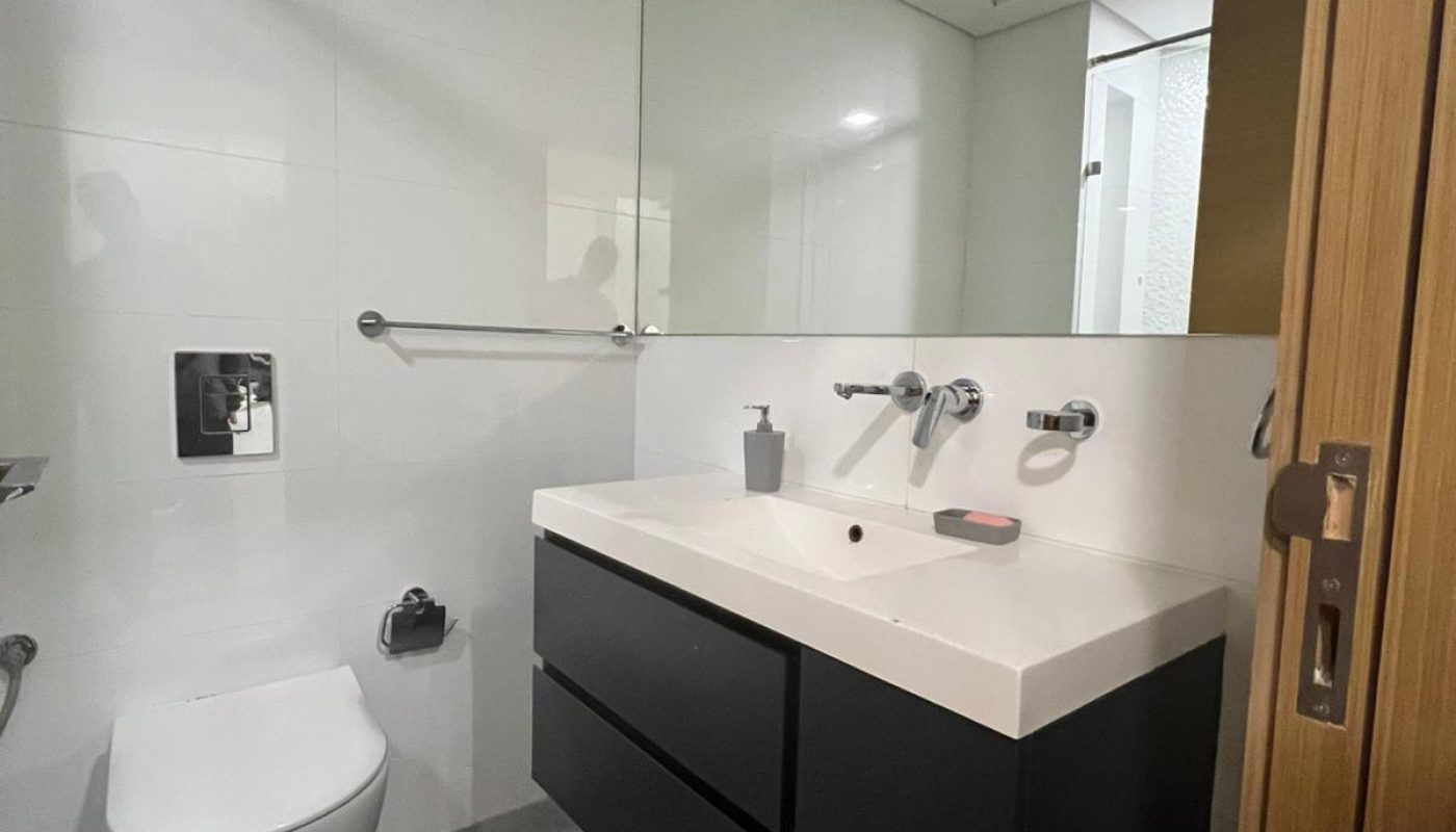 Modern bathroom with wall-mounted toilet, large mirror, black vanity with two drawers, and soap dispenser on the sink. Trash bin is on the gray tiled floor near the door.