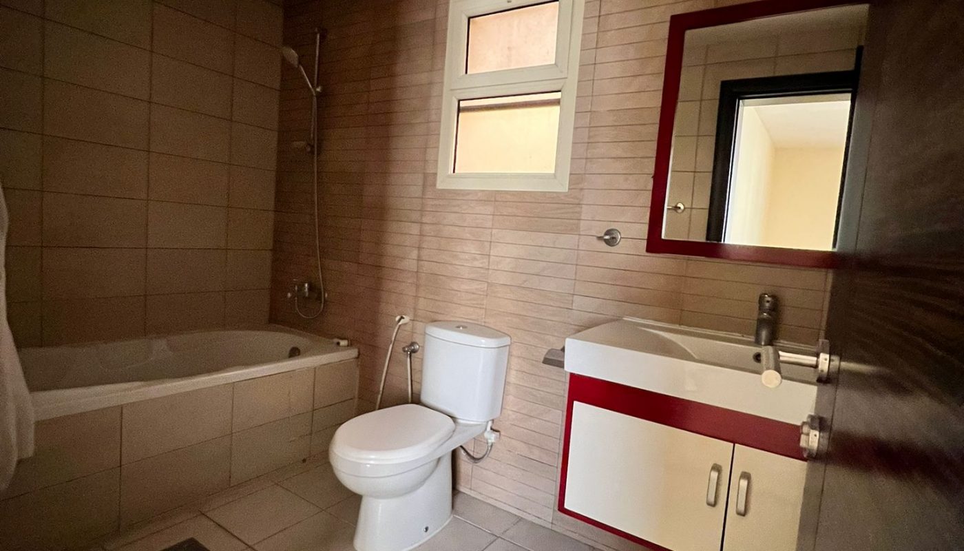 Bathroom with tiled floor and walls, a bathtub with shower, a toilet, a sink with a mirror, and a small window above the toilet.