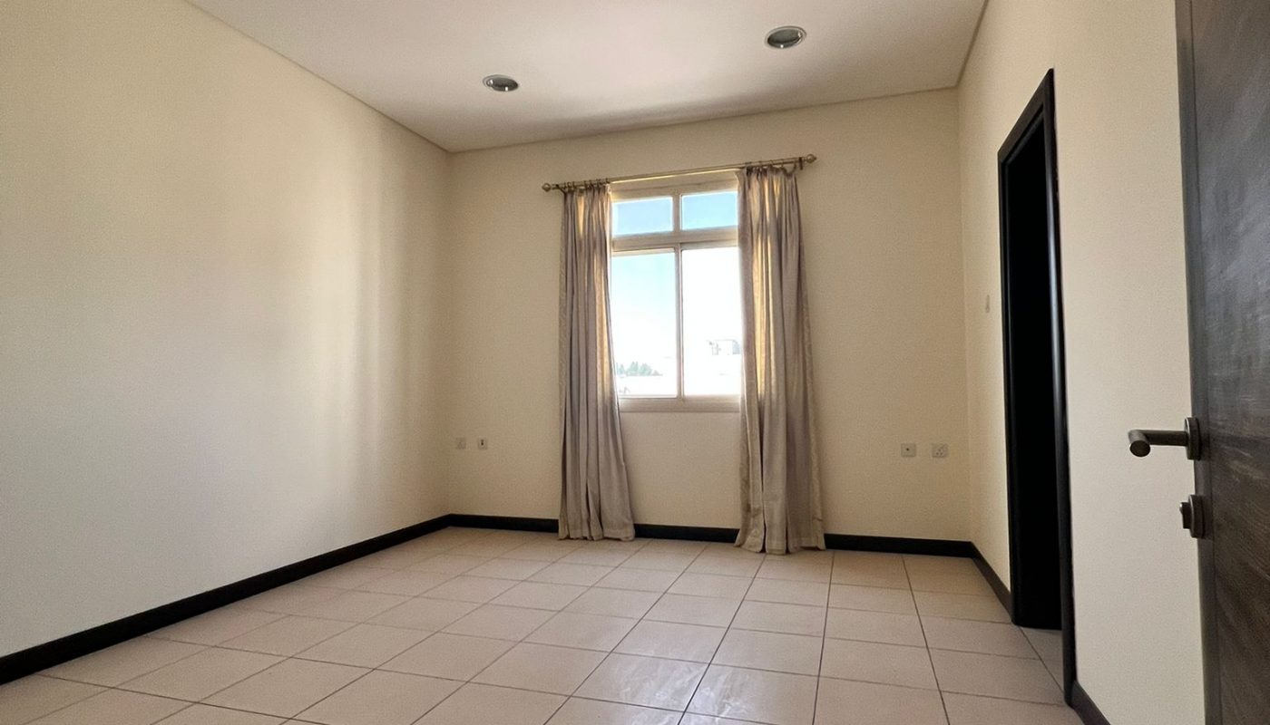 Empty room with cream-colored walls, tiled floor, a window with beige curtains, ceiling lights, and an open door on the right.
