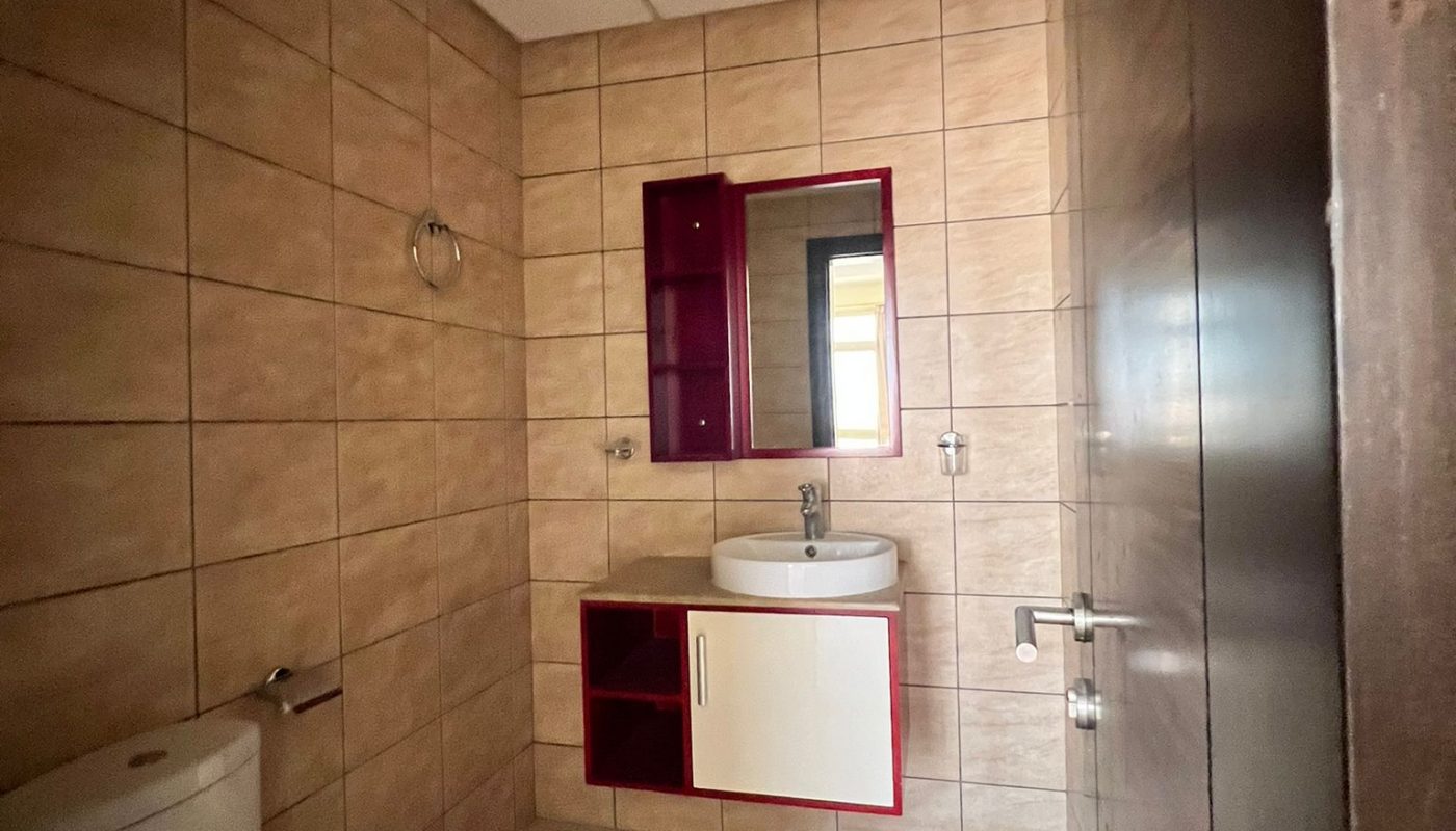 Small bathroom with beige tiled walls and floor, a toilet, a sink with a cabinet and a mirror above it, and a towel ring on the left wall.