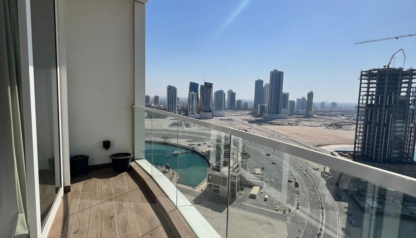 A high-rise balcony with a glass railing overlooks a cityscape featuring several tall buildings, wide roads, a body of water, and ongoing construction under a clear blue sky.