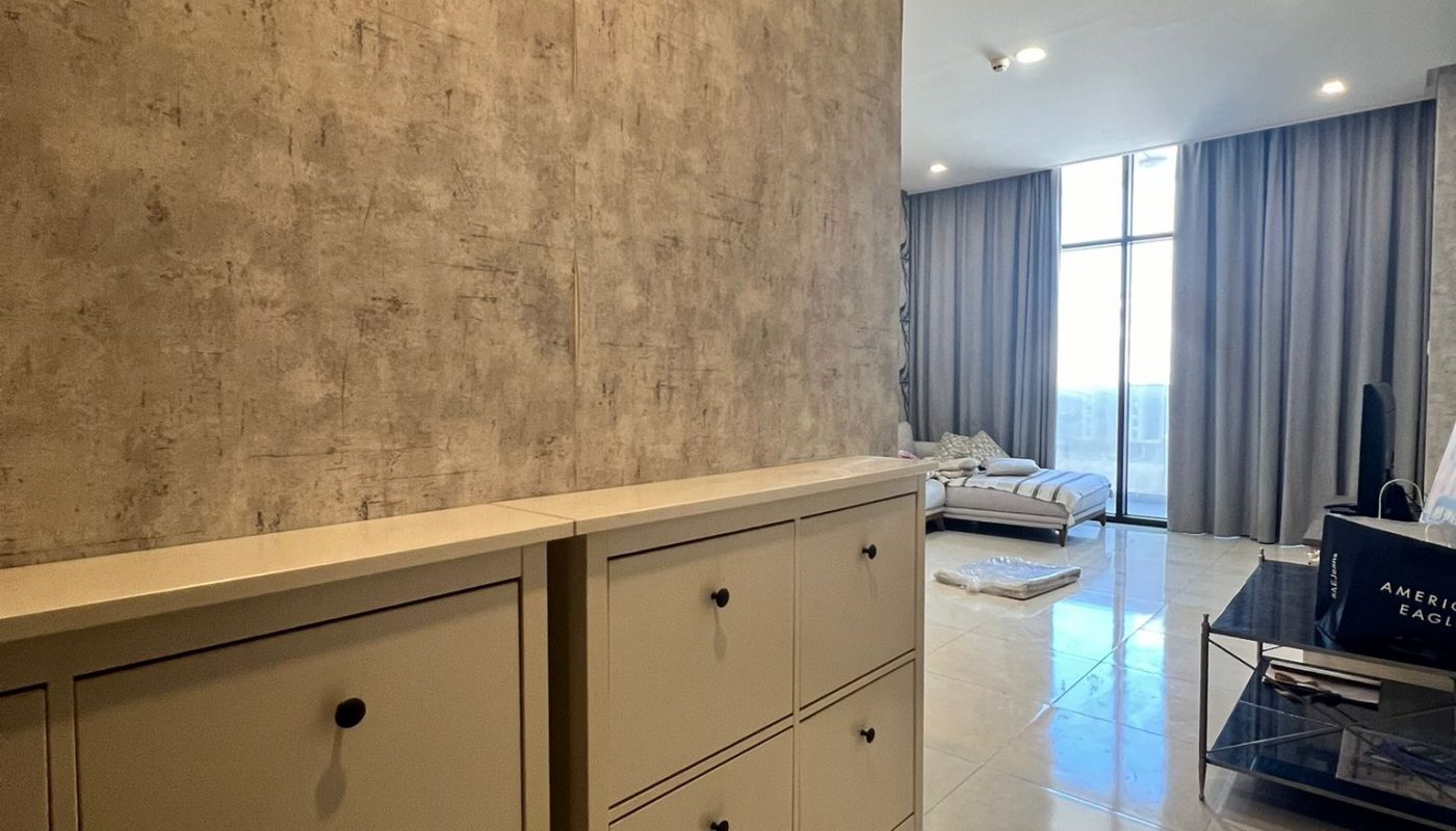 A modern, spacious living area with tiled floors, a textured wall, cream cabinets, large windows, and a couch near floor-to-ceiling curtains.