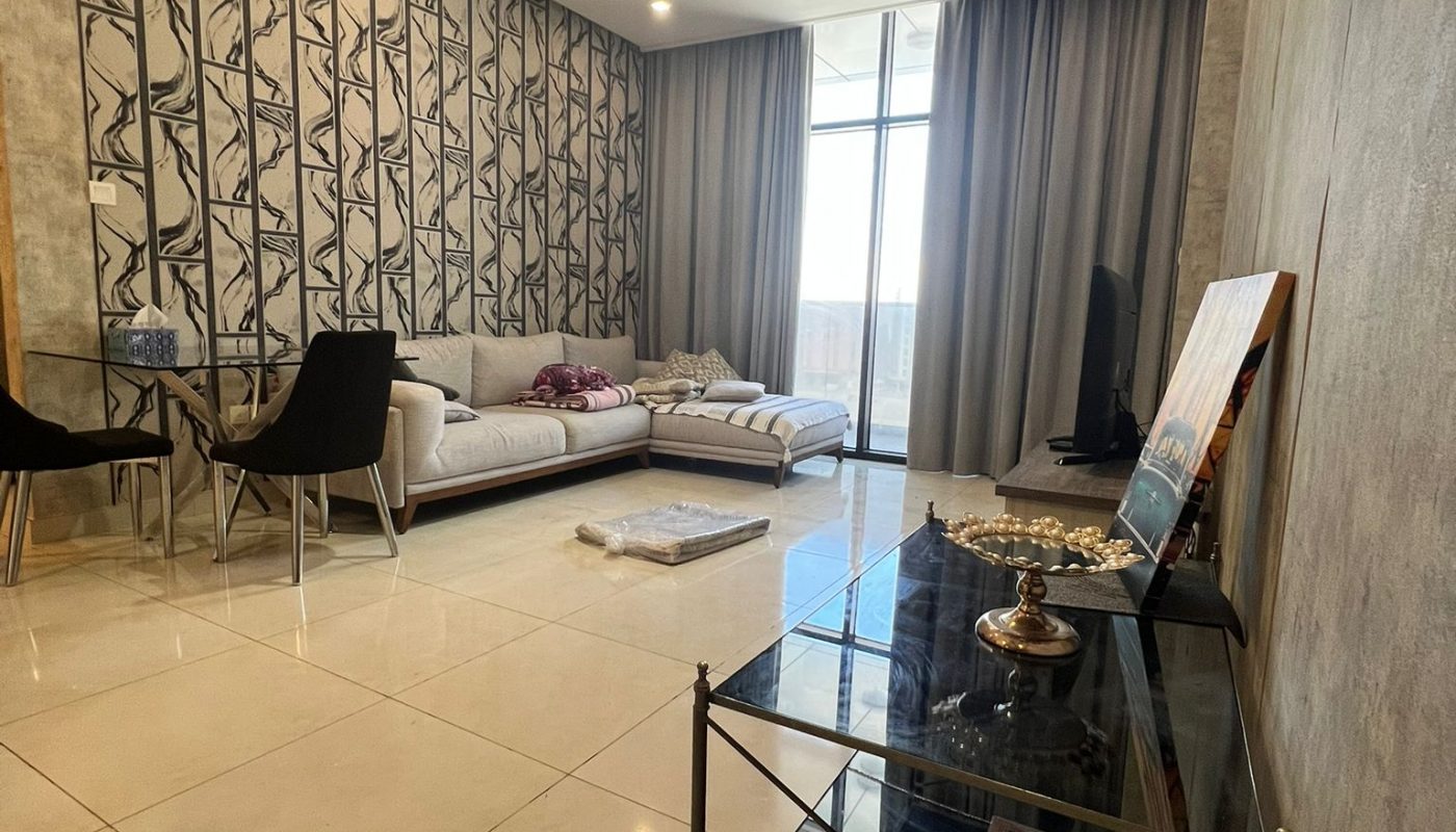 Modern living room with a white sectional sofa, patterned feature wall, glass table, chairs, and large window covered by curtains. A decorative shelf and wall clock are in the foreground.