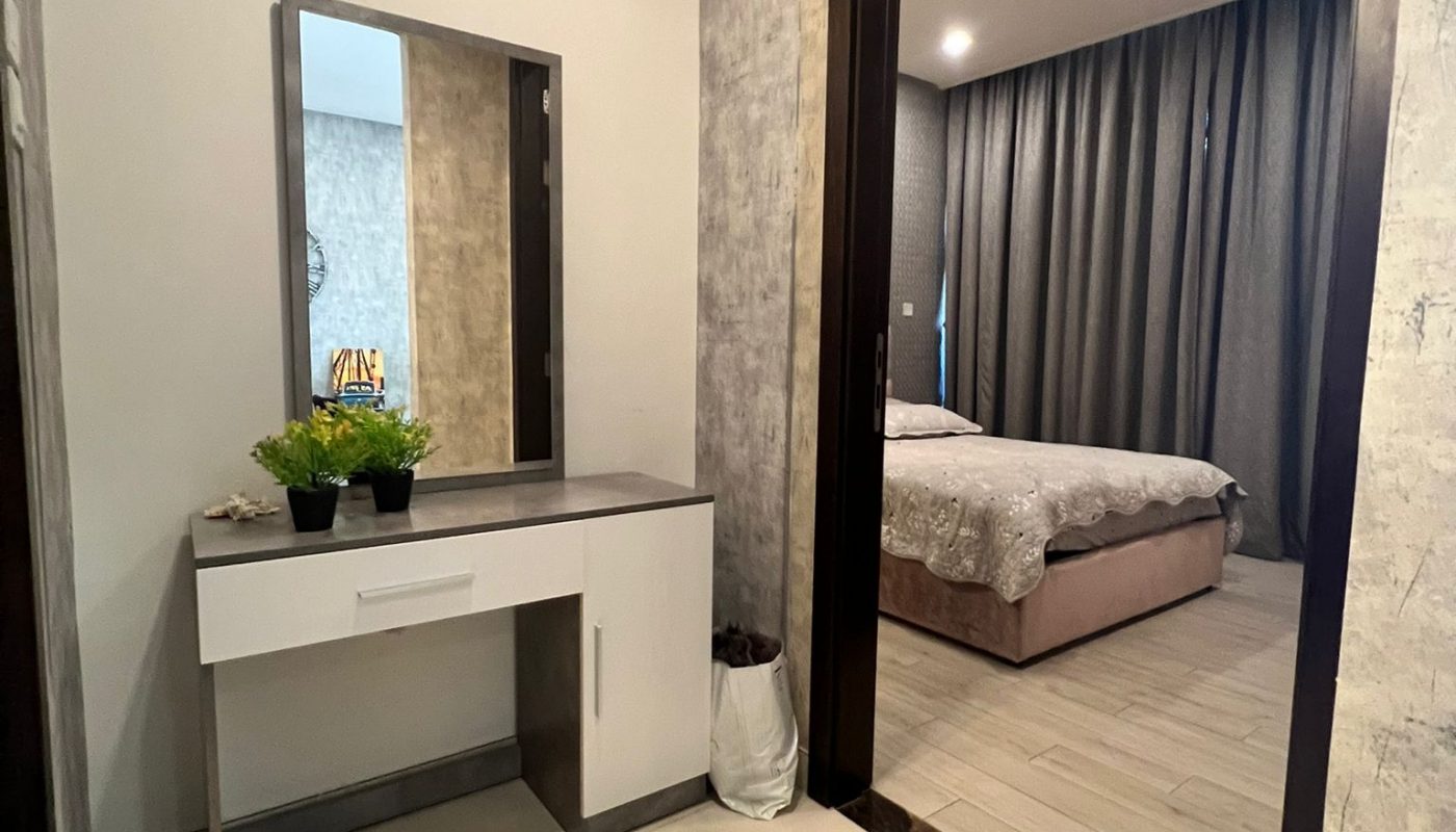 A small vanity with a plant and mirror stands in a tiled hallway next to an open doorway leading to a bedroom with a bed and gray curtains.