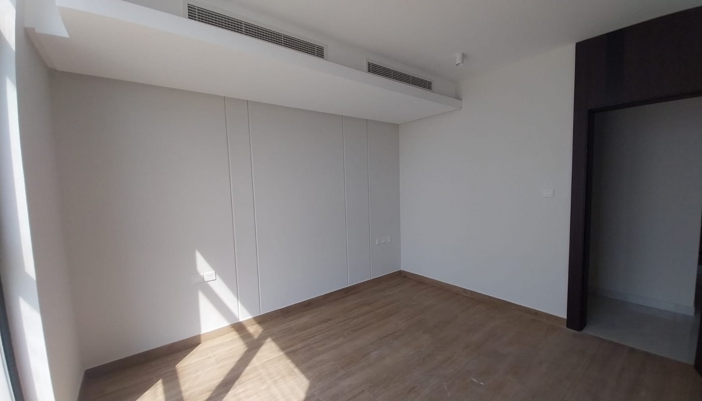 Empty room with light-colored walls, wooden floor, ceiling vents, and sunlight streaming through a window; doorway leads to another area.
