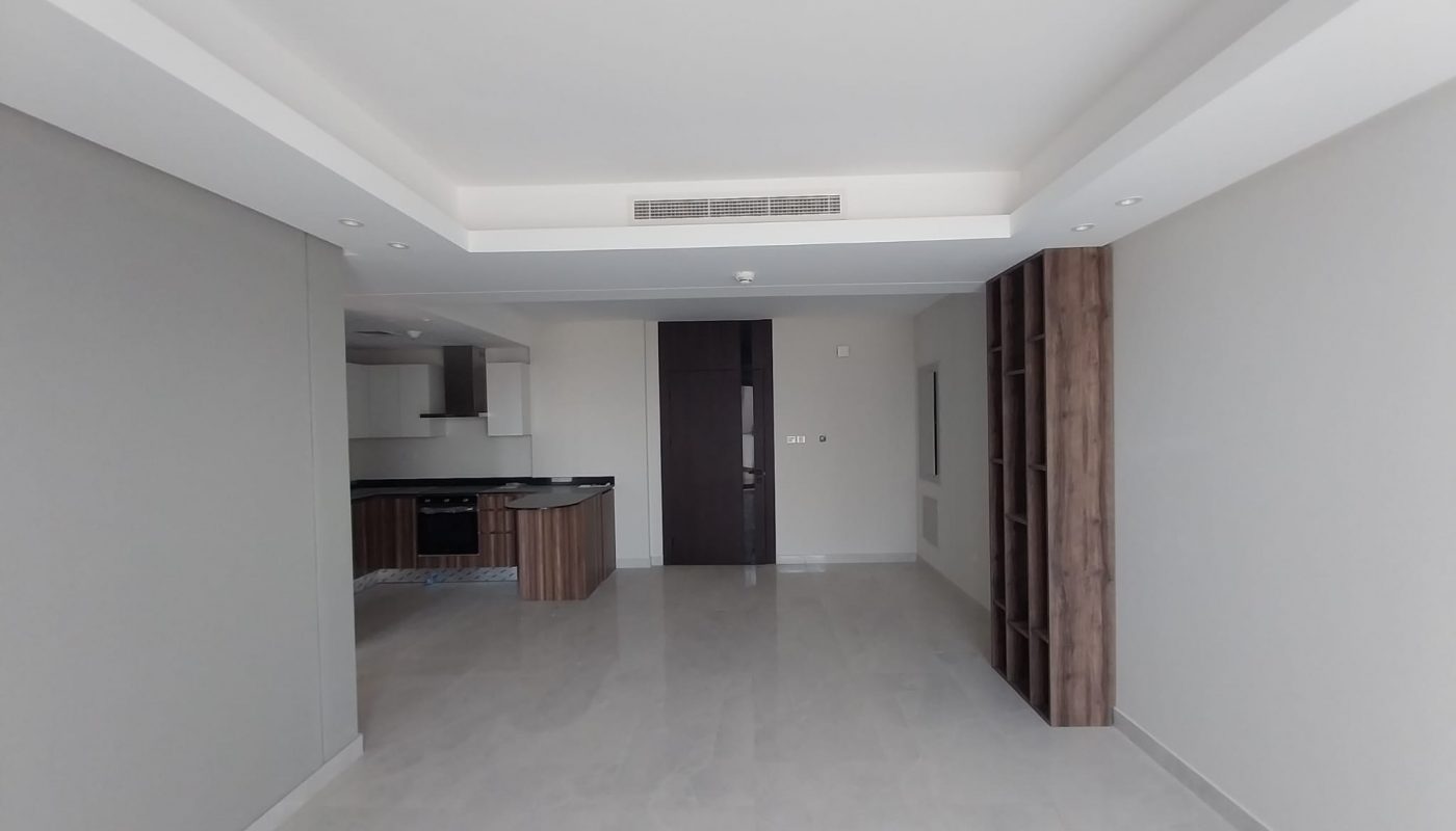 A modern, unfurnished open-plan apartment with a kitchen area, tiled floor, built-in shelves, and recessed ceiling lights.