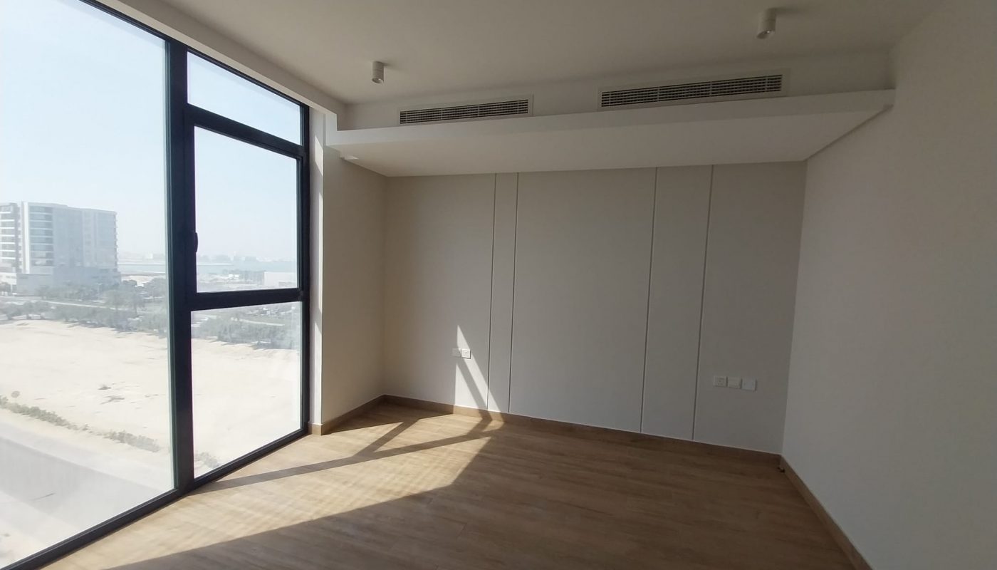 Empty room with large windows, beige walls, wooden floor, and ceiling air vents. Sunlight is casting a shadow on the floor. Urban buildings are visible outside.