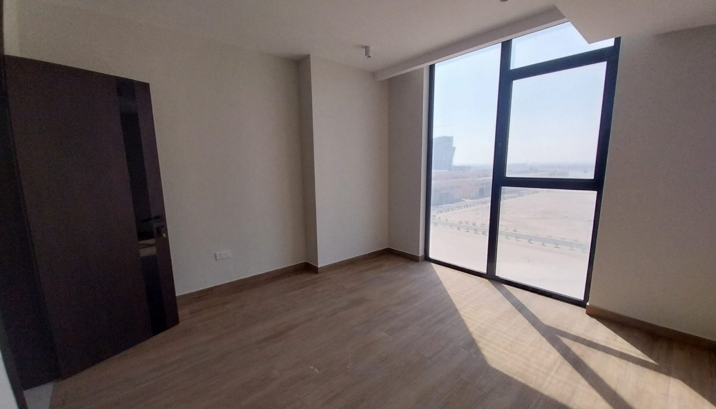 Empty room with large floor-to-ceiling windows, wood-style flooring, white walls, and ceiling light fixtures, with sunlight streaming in through the window.