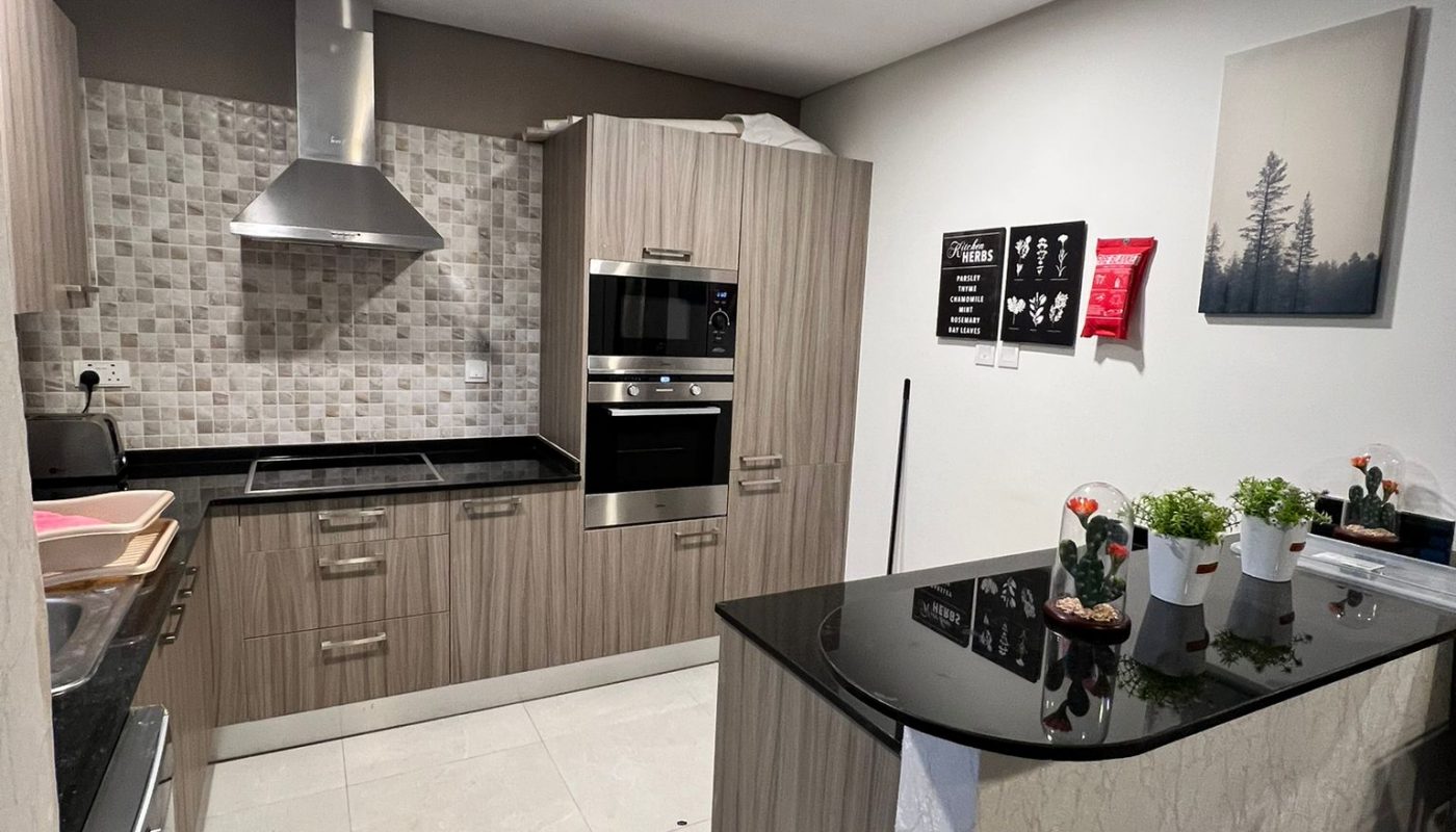 Modern kitchen with wood cabinets, black countertops, stainless steel appliances, a cooker hood, potted plants, and wall decor under bright ceiling lights.