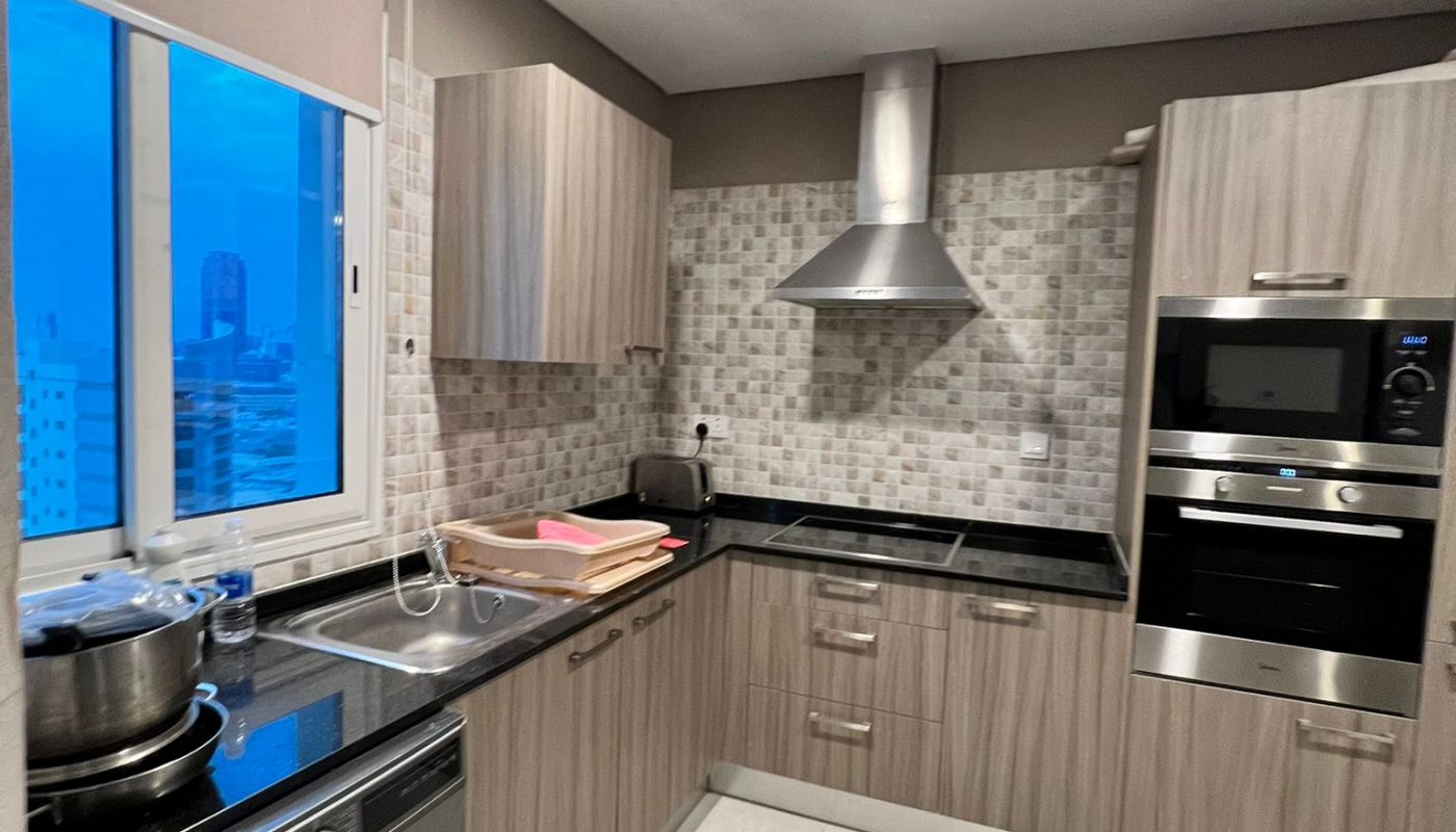 Modern kitchen with wood cabinets, a built-in oven, washing machine, stainless steel range hood, countertop appliances, and a window showing a cityscape at dusk.
