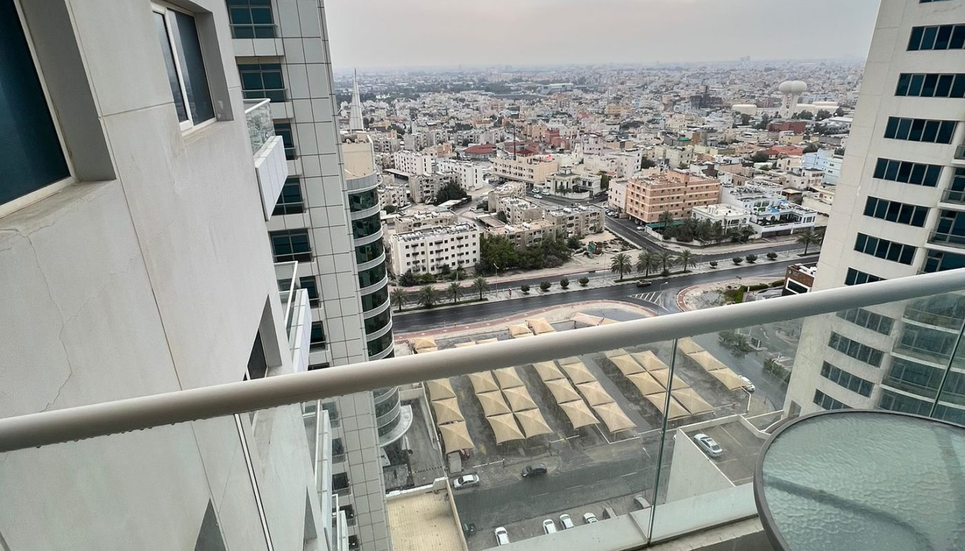 View from a high-rise balcony showing city buildings, a glass railing, a round glass table, and overcast sky in the background.