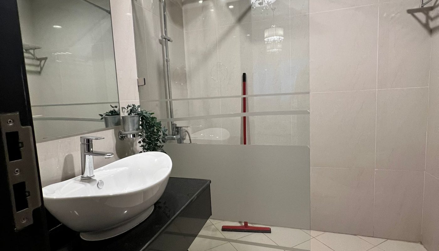 Modern bathroom with a glass-enclosed shower, round sink on a black counter, wall mirror, mop, towel rack, and a small plant. Beige tiles cover the walls and floor.