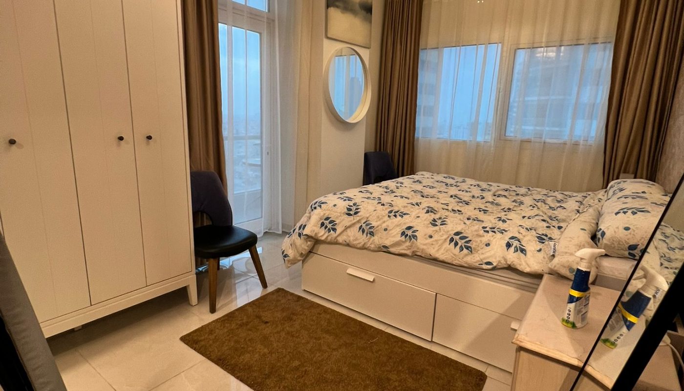 A neatly arranged bedroom with a double bed, white wardrobe, dresser, mirror, two chairs, and two ceiling lights, featuring beige curtains and tiled floor.