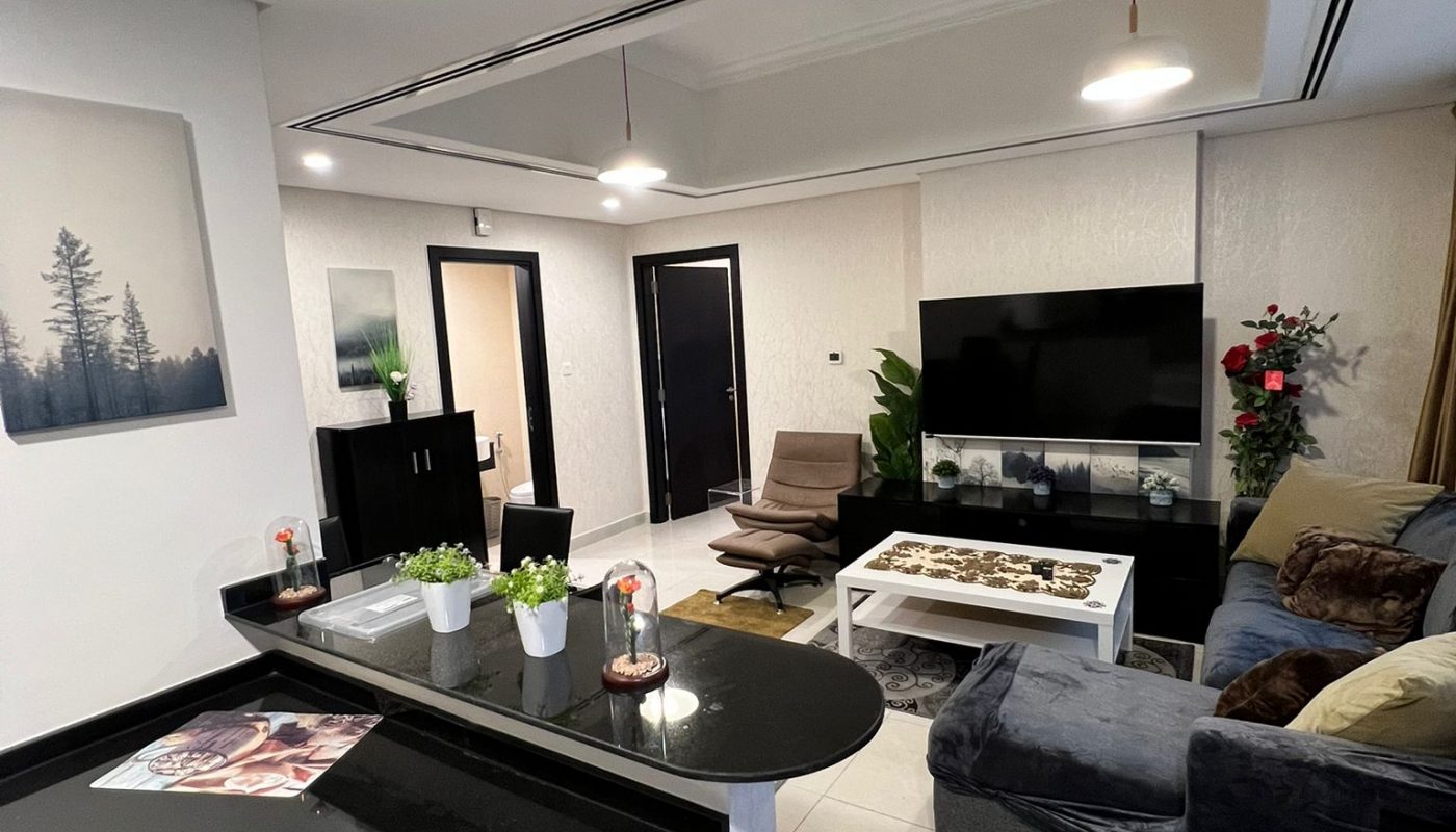 Modern living room with a sectional sofa, TV, potted plants, and kitchen counter; neutral colors and minimalistic decor.